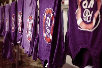 A line of purple shirts are hanging, each featuring a circular logo with the letters 'R' and 'P' surrounded by a red and white design. The shirts appear to be drying outside.