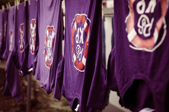 A line of purple shirts are hanging, each featuring a circular logo with the letters 'R' and 'P' surrounded by a red and white design. The shirts appear to be drying outside.
