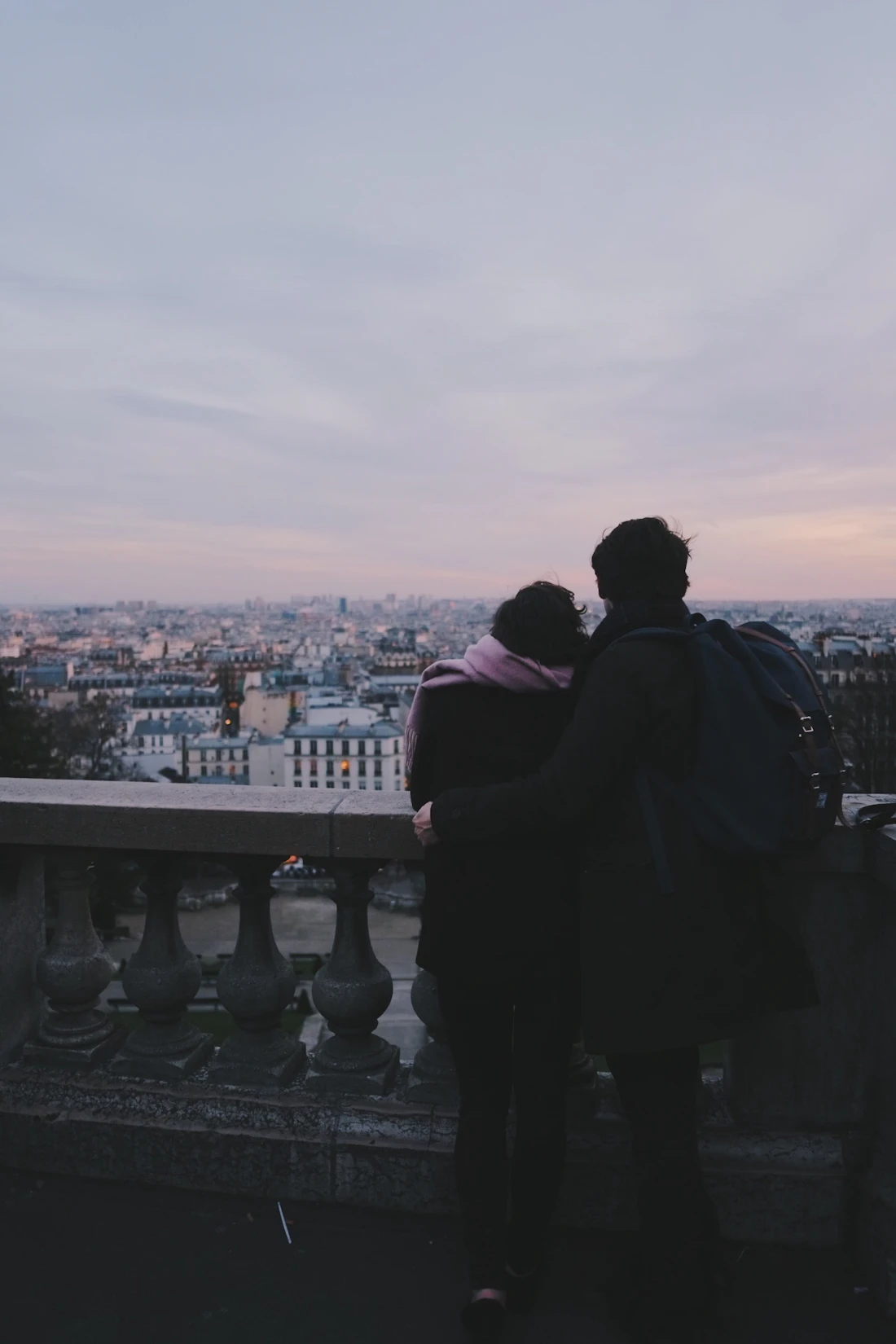 Two people standing arm in arm at a stone balustrade in Montmartre, looking out over the Paris rooftops at dusk.