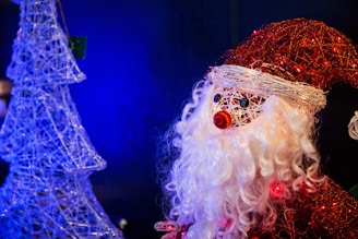 A decorative figure resembling Santa Claus made from woven materials features prominently, sporting a red hat and white beard. To the left, a wireframe Christmas tree structure is illuminated against a blue backdrop, creating a festive ambiance.