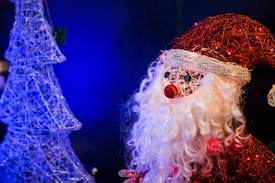 A decorative figure resembling Santa Claus made from woven materials features prominently, sporting a red hat and white beard. To the left, a wireframe Christmas tree structure is illuminated against a blue backdrop, creating a festive ambiance.