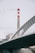 A tall chimney with red and white stripes emits a small amount of smoke in an industrial urban area. The foreground features a large modern bridge with a lattice structure. Several buildings, including one with a red and white striped pattern, can be seen in the background.