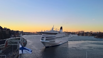 A large white cruise ship sails through a calm waterway during sunset, with a city skyline featuring historic buildings and cranes visible in the background. A flag with a blue cross flies in the foreground on a dock.
