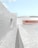 A minimalist architectural scene features smooth, curved concrete structures under a clear blue sky. The design uses pale colors, such as soft pink and white, creating a serene and modern feel. The ground is paved with light-colored stone tiles.