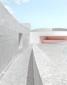 A minimalist architectural scene features smooth, curved concrete structures under a clear blue sky. The design uses pale colors, such as soft pink and white, creating a serene and modern feel. The ground is paved with light-colored stone tiles.