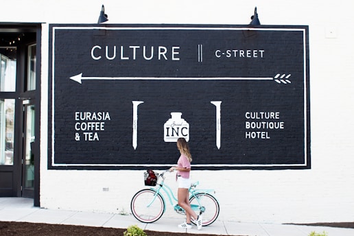 A woman wearing a pink top and white shorts is standing next to a teal bicycle with a basket, in front of a wall with a large black and white mural. The mural displays text that includes 'CULTURE', 'C-STREET', and names of businesses like 'EURASIA COFFEE & TEA' and 'CULTURE BOUTIQUE HOTEL'.
