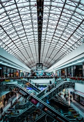 A modern shopping mall interior featuring a large, geometric glass ceiling. Multiple levels are connected by intersecting escalators. The area is bustling with people shopping and passing by various retail stores lining the sides.
