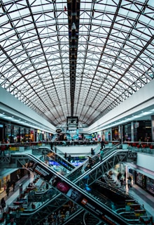 A modern shopping mall interior featuring a large, geometric glass ceiling. Multiple levels are connected by intersecting escalators. The area is bustling with people shopping and passing by various retail stores lining the sides.