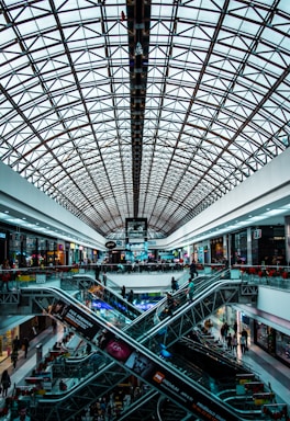 A modern shopping mall interior featuring a large, geometric glass ceiling. Multiple levels are connected by intersecting escalators. The area is bustling with people shopping and passing by various retail stores lining the sides.