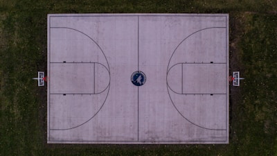 An overhead view of an outdoor basketball court with a concrete surface surrounded by greenery. The court is marked with standard basketball lines, featuring two hoops with orange rims at opposite ends.