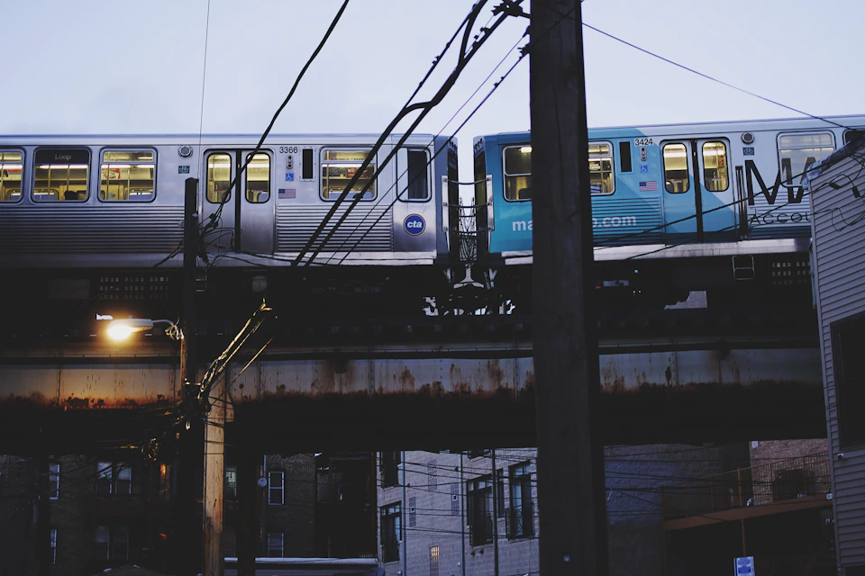 The image depicts a train crossing over an elevated railway track, with two trains positioned side by side on parallel tracks. The first train is silver and blue, while the second train is white and blue. Both trains are moving towards each other, creating a sense of motion in the scene. In the background, there are buildings visible, suggesting that this location might be near an urban area or city center.