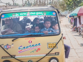 A crowded autorickshaw filled with children wearing school uniforms and a driver in the front on a street lined with market stalls under colorful umbrellas.