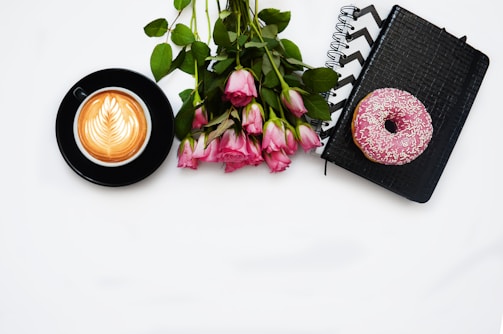 A black cup containing coffee with intricate latte art sits beside a bouquet of pink roses on a white surface. Nearby, a pink frosted donut with sprinkles rests on top of a black textured notebook with a zigzag design.
