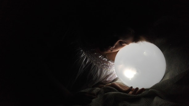 A person is lying peacefully in the dark, with their head resting near a glowing orb, creating a soft illumination. The setting appears tranquil, with a focus on the gentle light and soft shadows cast around the figure.