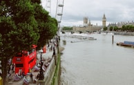 A riverside view featuring the iconic London Eye and the Palace of Westminster. The scene includes a red double-decker bus parked near the walkway lined with trees and colorful deck chairs. People are walking along the riverside promenade, and the river is busy with boats.
