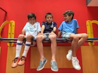Three young boys are sitting on a playground structure. Each boy has a cheerful expression and is dressed casually, with sneakers and shorts. The background is vibrant red with yellow accents, and they appear to be enjoying their time together.