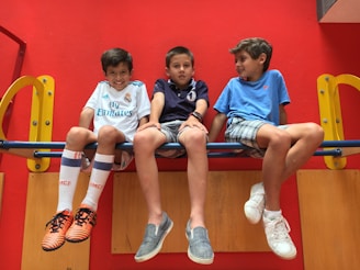 Three young boys are sitting on a playground structure. Each boy has a cheerful expression and is dressed casually, with sneakers and shorts. The background is vibrant red with yellow accents, and they appear to be enjoying their time together.