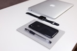 A neatly arranged collection of Apple products and office supplies on a white desk. A closed silver MacBook, a black Apple Watch with a black strap, a black iPhone, and a black pen are all positioned in an orderly manner. The pen and iPhone are placed on top of a closed grey notebook.