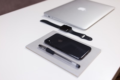 A neatly arranged collection of Apple products and office supplies on a white desk. A closed silver MacBook, a black Apple Watch with a black strap, a black iPhone, and a black pen are all positioned in an orderly manner. The pen and iPhone are placed on top of a closed grey notebook.