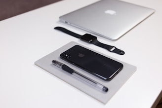 A neatly arranged collection of Apple products and office supplies on a white desk. A closed silver MacBook, a black Apple Watch with a black strap, a black iPhone, and a black pen are all positioned in an orderly manner. The pen and iPhone are placed on top of a closed grey notebook.