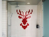 A shower area with white tiles featuring a pixelated red stag design on the back wall. The shower fixtures include a modern square showerhead and a handheld shower wand. The space is enclosed by a glass door and has a minimalist style.