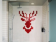 A shower area with white tiles featuring a pixelated red stag design on the back wall. The shower fixtures include a modern square showerhead and a handheld shower wand. The space is enclosed by a glass door and has a minimalist style.