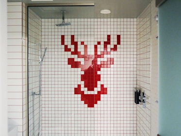 A shower area with white tiles featuring a pixelated red stag design on the back wall. The shower fixtures include a modern square showerhead and a handheld shower wand. The space is enclosed by a glass door and has a minimalist style.
