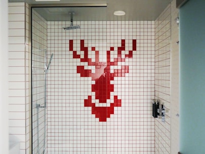 A shower area with white tiles featuring a pixelated red stag design on the back wall. The shower fixtures include a modern square showerhead and a handheld shower wand. The space is enclosed by a glass door and has a minimalist style.