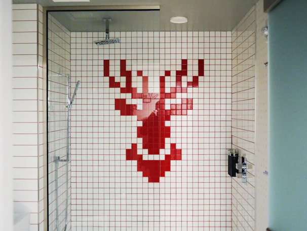 A shower area with white tiles featuring a pixelated red stag design on the back wall. The shower fixtures include a modern square showerhead and a handheld shower wand. The space is enclosed by a glass door and has a minimalist style.