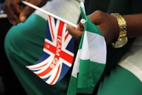 A person wearing a gold watch holds two small flags. One flag is the Union Jack with the words 'TEAM GB' printed on it, and the other is a green and white flag, possibly representing Nigeria.