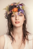 A person with wet hair wearing a floral crown. The crown includes vibrant flowers such as purple irises, yellow blooms, orange petals, and small pink buds. The individual is dressed in a white sleeveless top, with a neutral expression and a slightly open mouth.