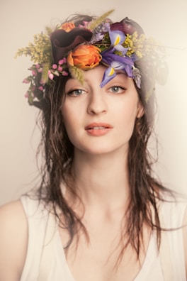 A person with wet hair wearing a floral crown. The crown includes vibrant flowers such as purple irises, yellow blooms, orange petals, and small pink buds. The individual is dressed in a white sleeveless top, with a neutral expression and a slightly open mouth.