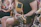 A person wearing shorts and a dark t-shirt is playing an acoustic guitar while seated on a floral-patterned armchair. The focus is on the guitar and hands. Nearby, a woman in a colorful patterned dress is partially visible, suggesting a relaxed, social setting.