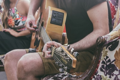 A person wearing shorts and a dark t-shirt is playing an acoustic guitar while seated on a floral-patterned armchair. The focus is on the guitar and hands. Nearby, a woman in a colorful patterned dress is partially visible, suggesting a relaxed, social setting.
