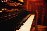 A close-up view of a piano keyboard with a blurred, warm-toned background. The piano keys are well-lit, creating a contrast against the darker surroundings. Reflections and soft lights add to the atmosphere, suggesting a cozy, intimate setting.