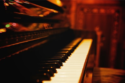 A close-up view of a piano keyboard with a blurred, warm-toned background. The piano keys are well-lit, creating a contrast against the darker surroundings. Reflections and soft lights add to the atmosphere, suggesting a cozy, intimate setting.