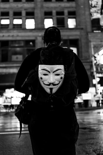 grayscale photography of person walking guy fawkes mask backpack
