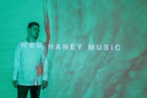 A person wearing a light-colored sweater stands against a textured, teal background. The words 'WESHANEY MUSIC' are displayed across the image in bold, white letters. There is a reddish vertical line running down beside the person, blending with the background.