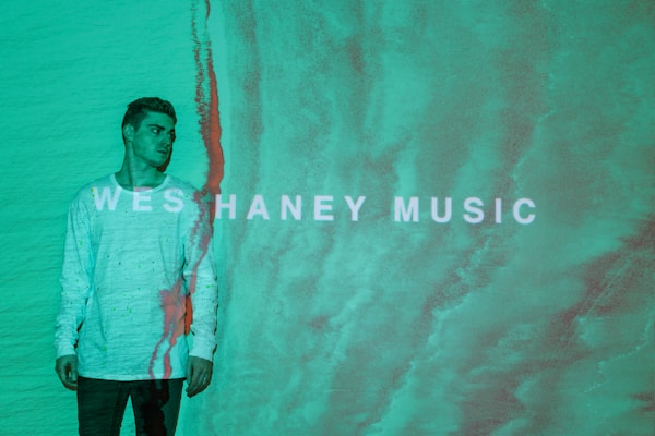 A person wearing a light-colored sweater stands against a textured, teal background. The words 'WESHANEY MUSIC' are displayed across the image in bold, white letters. There is a reddish vertical line running down beside the person, blending with the background.
