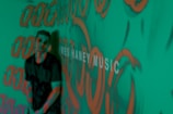 A person stands in front of a green background covered with orange graffiti-like patterns. The words 'MUSIC' are partially visible, adding an artistic and urban vibe to the scene.