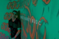 A person stands in front of a green background covered with orange graffiti-like patterns. The words 'MUSIC' are partially visible, adding an artistic and urban vibe to the scene.