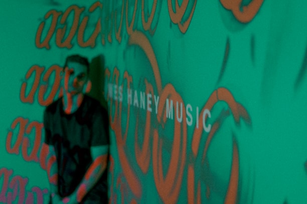 A person stands in front of a green background covered with orange graffiti-like patterns. The words 'MUSIC' are partially visible, adding an artistic and urban vibe to the scene.