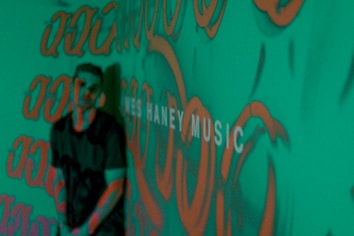 A person stands in front of a green background covered with orange graffiti-like patterns. The words 'MUSIC' are partially visible, adding an artistic and urban vibe to the scene.