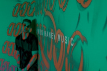 A person stands in front of a green background covered with orange graffiti-like patterns. The words 'MUSIC' are partially visible, adding an artistic and urban vibe to the scene.