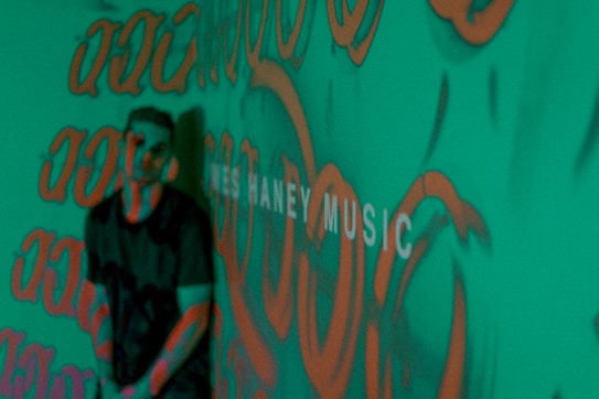 A person stands in front of a green background covered with orange graffiti-like patterns. The words 'MUSIC' are partially visible, adding an artistic and urban vibe to the scene.