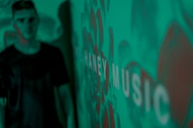 A blurred figure stands in front of a green background with a pattern and the word 'MUSIC' visible. The image has a pixelated digital display appearance.