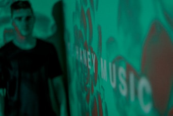 A blurred figure stands in front of a green background with a pattern and the word 'MUSIC' visible. The image has a pixelated digital display appearance.