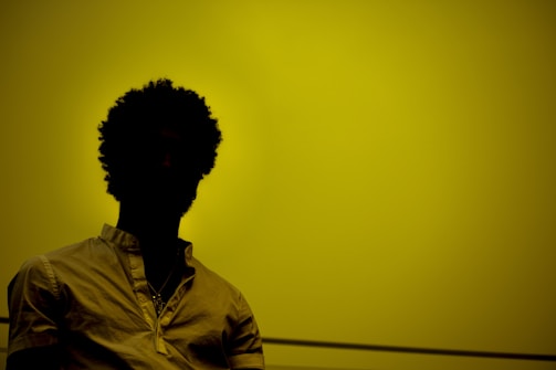 A silhouette of a person with curly hair against a yellow background, wearing a light-colored shirt.