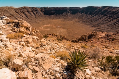 A large, barren impact crater surrounded by rocky and rugged terrain. The landscape is arid with sparse vegetation, including a noticeable agave plant in the foreground. The sky above is clear and blue, providing a stark contrast to the earthy tones below.