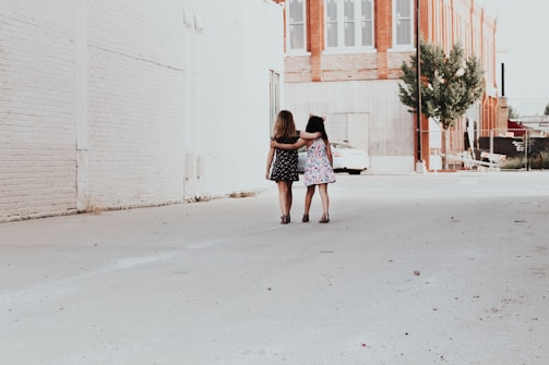 Two girls are walking down a deserted alleyway with their arms around each other. The buildings are primarily made of red brick and white painted walls, with some trees and a parked car further down the alley.